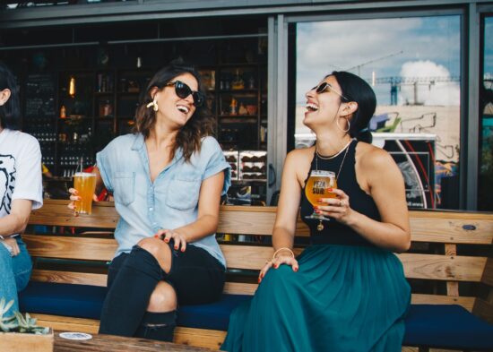 Two women laughing and drinking beer outside a bar.