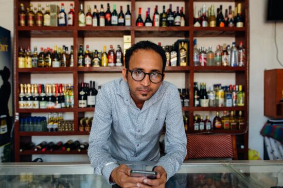 Man using a phone in front of shelves of liquor bottles.