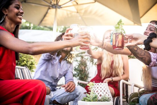 Friends toasting drinks outdoors under a patio umbrella.