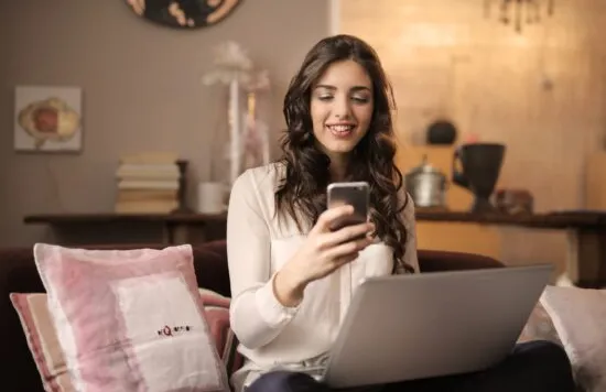 Woman using a phone and laptop on a couch.