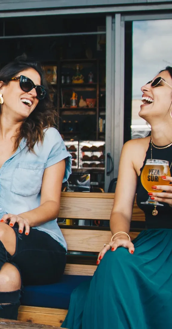 Two women sitting on a bench drinking beer in front of a bar